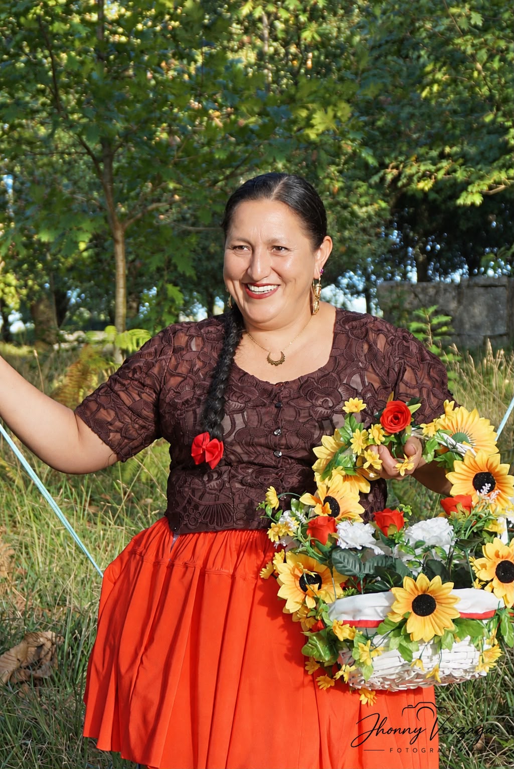 Una cholita sonriente con blusa marrón y pollera naranja lleva una canasta de girasoles