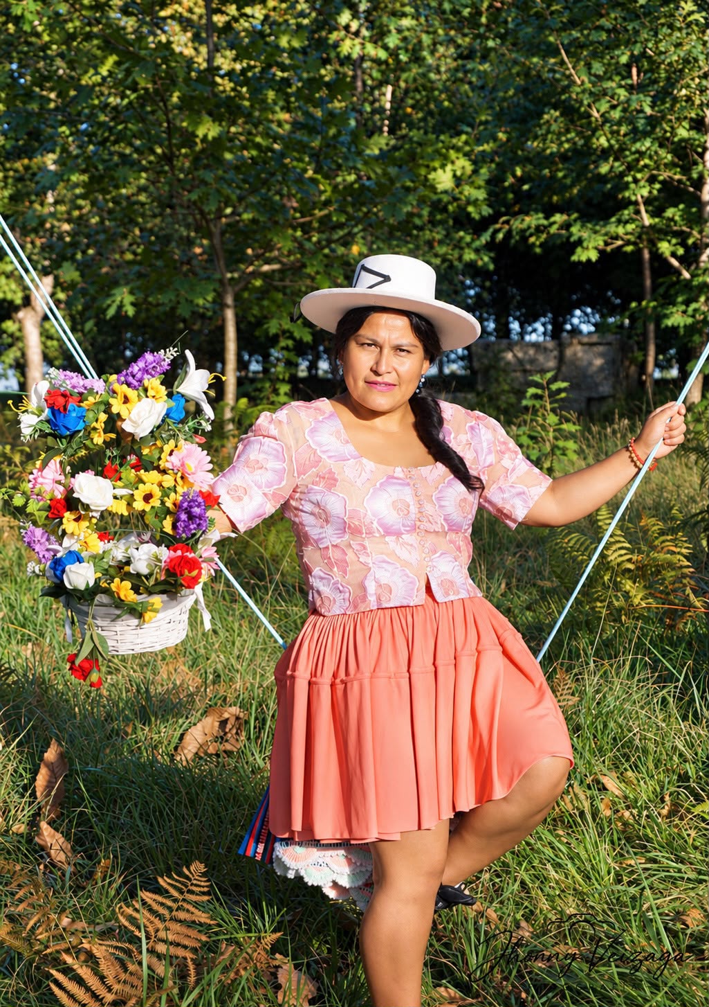 Una cholita con sombrero blanco y pollera salmón baila entre cuerdas de columpio con canastas de flores